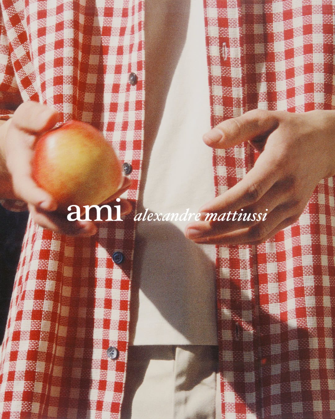 Close-up of a person wearing a red and white gingham shirt and holding an apple for Ami spring ad campaign.