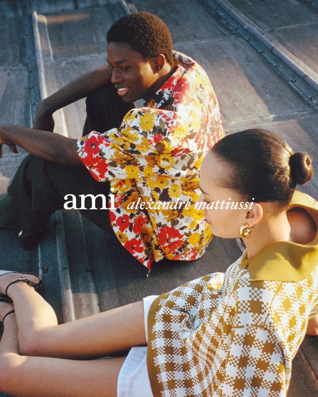 Two models seated on a rooftop during the Ami Spring 2025 ad campaign photographed by Crista Leonard in Paris