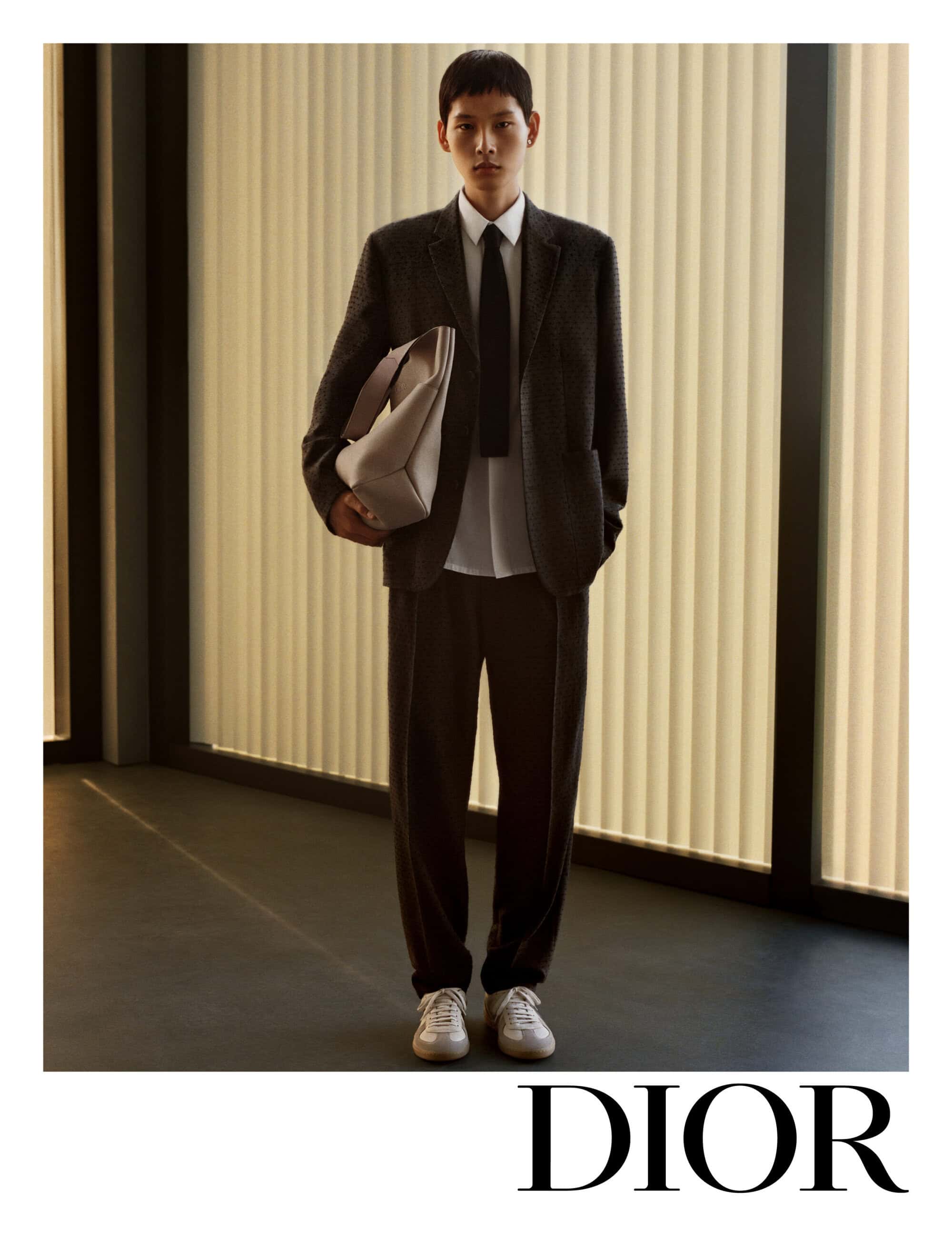 A male model wearing a dark suit and white shirt holds a large light-colored handbag against a vertically ribbed architectural background.