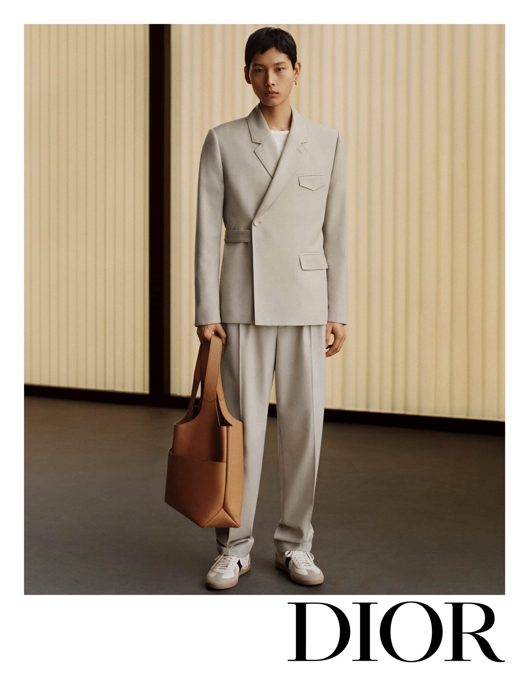 Model wearing a light grey suit and carrying a tan leather tote bag standing against a vertical paneled wall for Dior ad campaign