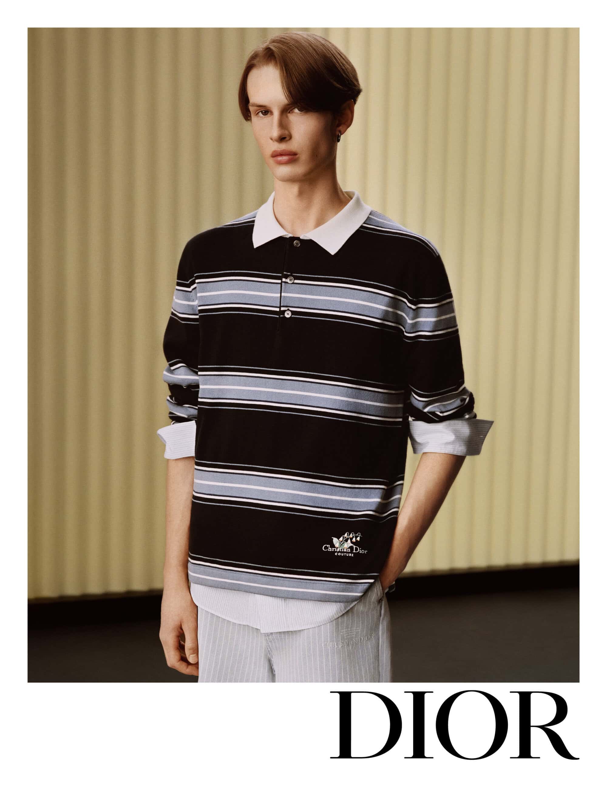 A male model wearing a black and blue striped polo shirt and white collared shirt against a ribbed vertical background.