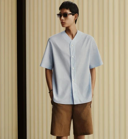 Model in sunglasses wearing a light blue short-sleeve shirt and brown shorts against a fluted panel background