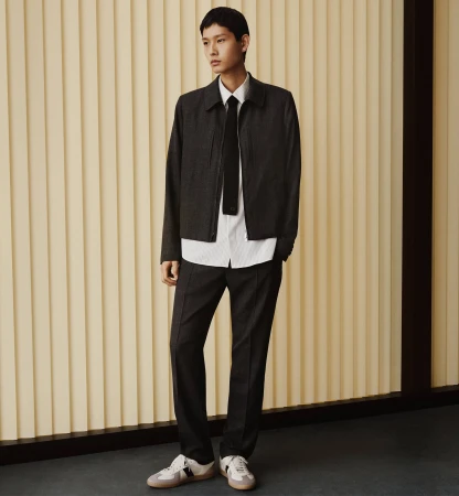 Model in a charcoal jacket, white shirt, and black tie standing against a vertically fluted cream wall for Dior.