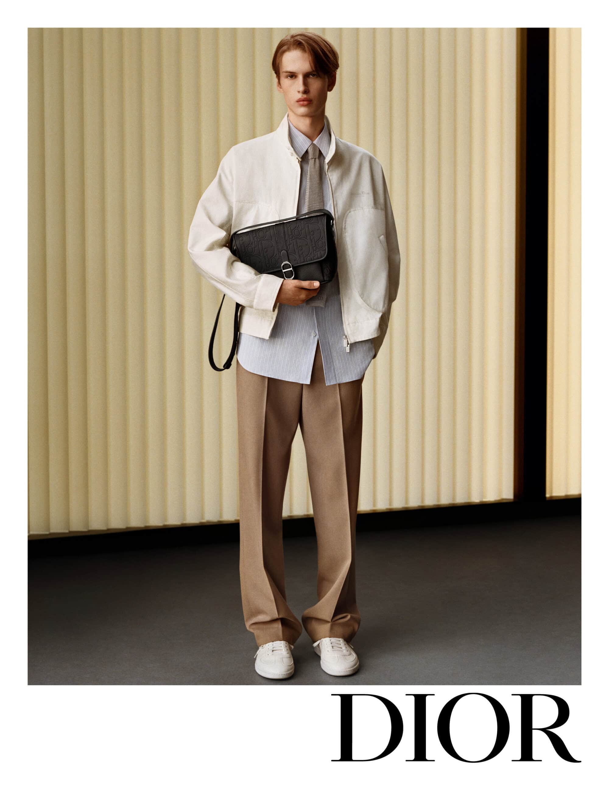 Model wearing a white jacket and beige trousers holding a black messenger bag in a minimalist interior setting