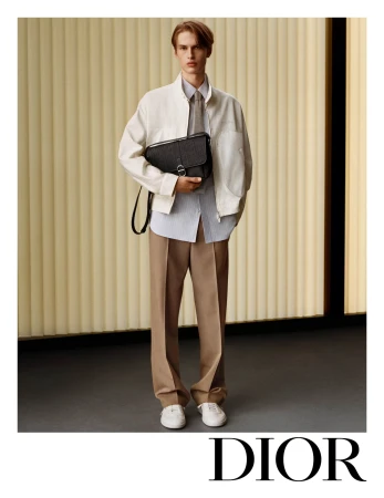 Model wearing a white jacket and beige trousers holding a black messenger bag in a minimalist interior setting