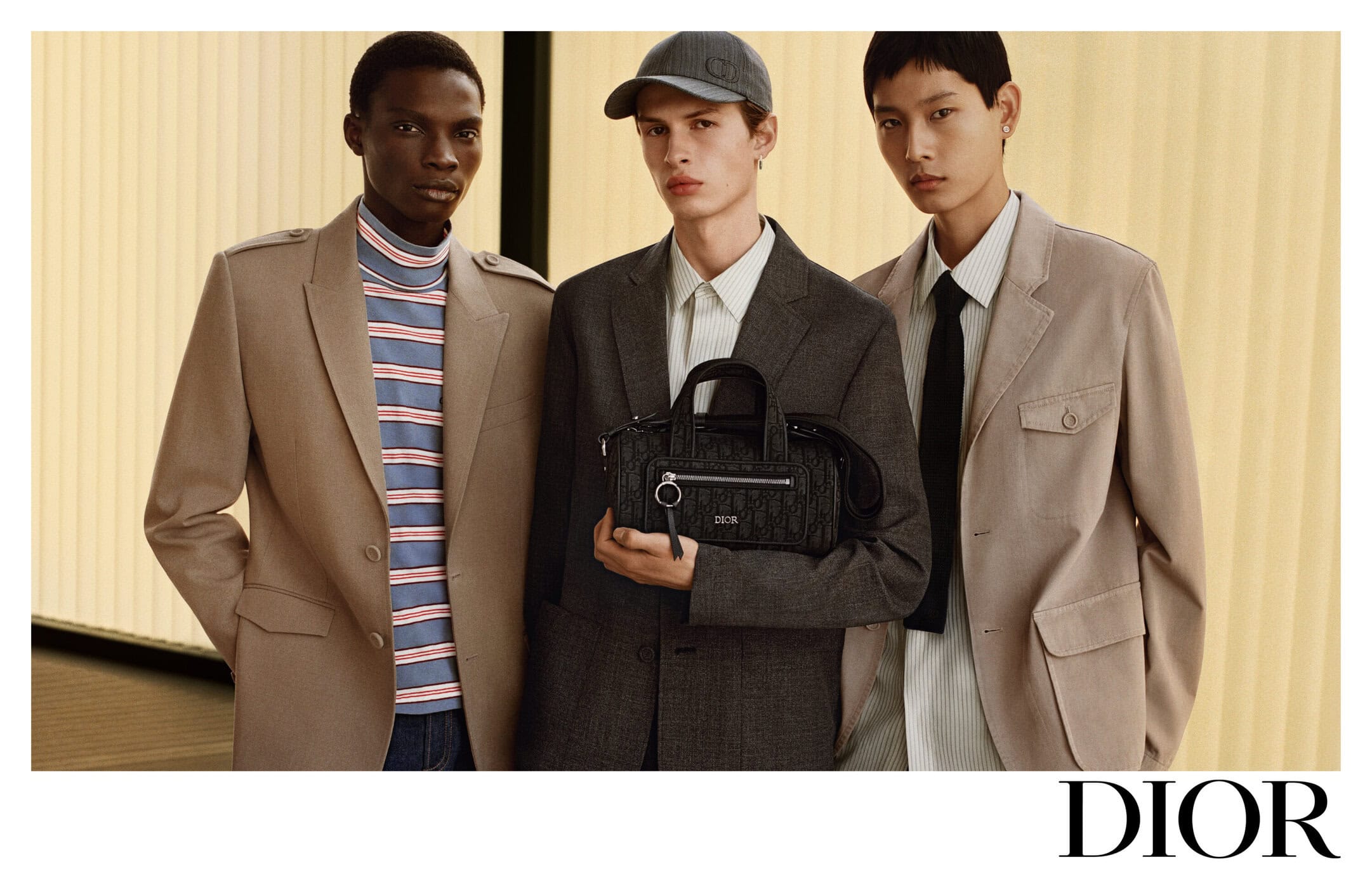 Three male models in tailored blazers posing against a slatted wall, with the central model holding a black monogram handbag.