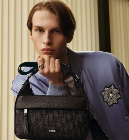 Close-up portrait of a male model holding a dark patterned shoulder bag wearing a blue bomber jacket for Dior Men's Fall 2025 ad campaign.