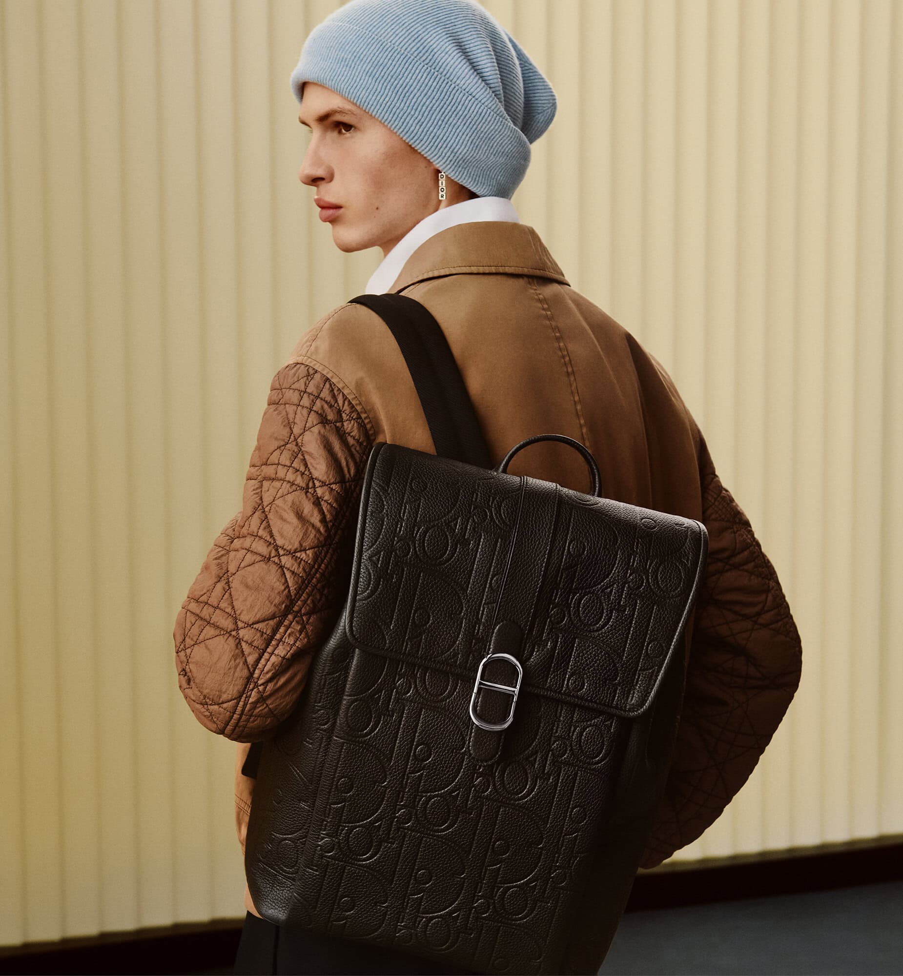 Model wearing a blue beanie and brown jacket carrying a black leather backpack against a vertical panel background.