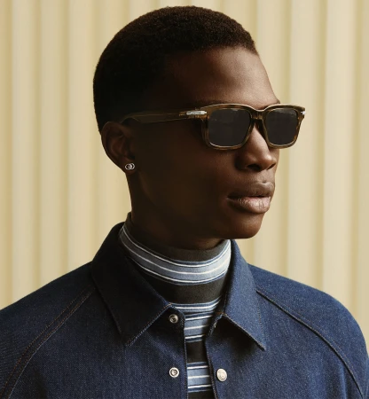 Man wearing sunglasses and a blue jacket against a textured wall for Dior Men's Fall 2025 campaign.