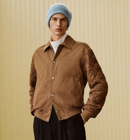 A male model wearing a light blue beanie, brown quilted jacket, and white turtleneck against a vertical ribbed background.