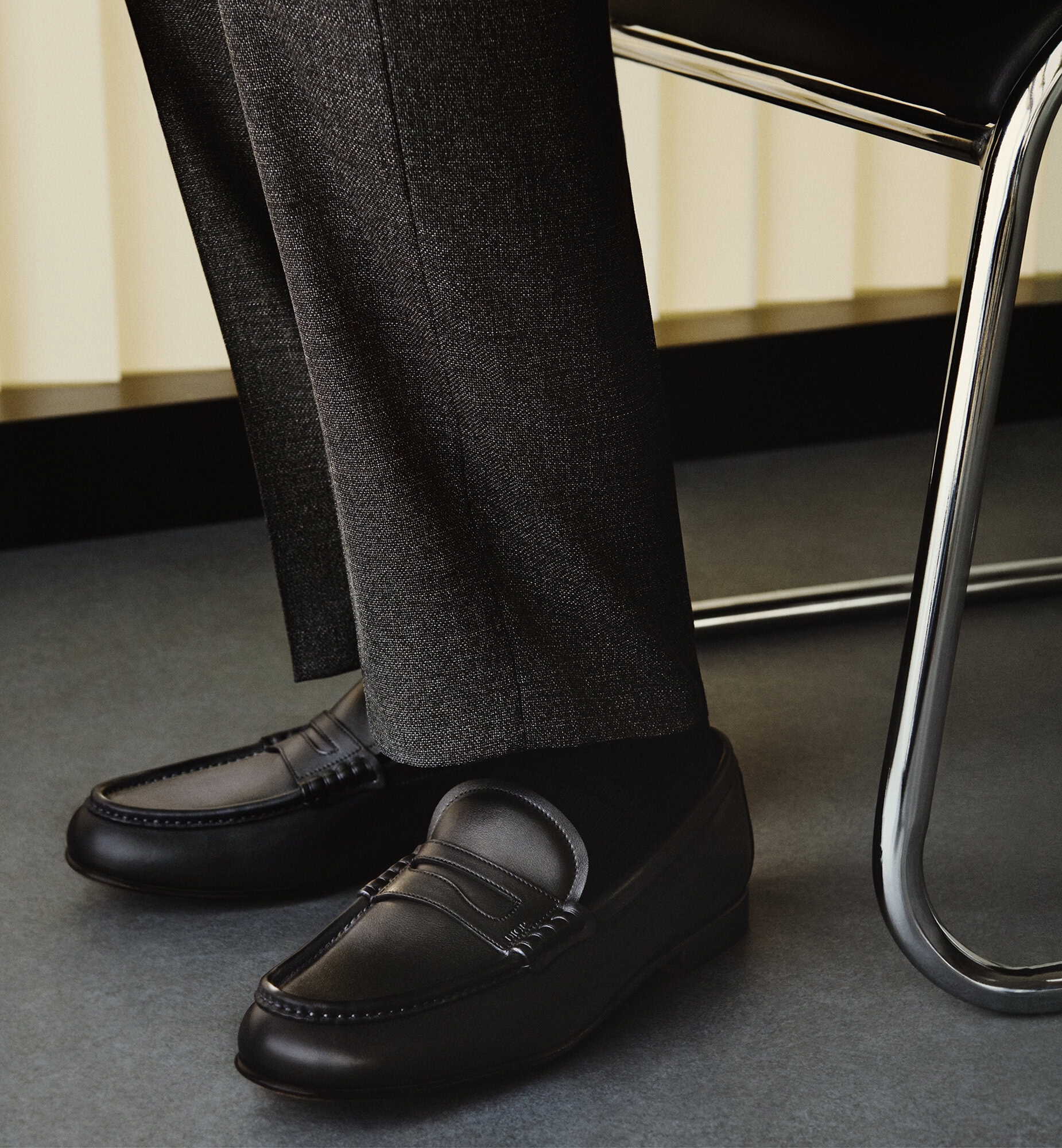 Close-up of black leather penny loafers worn with dark grey trousers next to a chrome and black chair