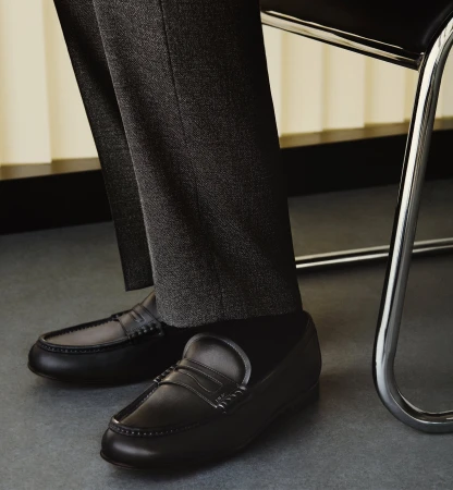 Close-up of black leather penny loafers worn with dark grey trousers next to a chrome and black chair