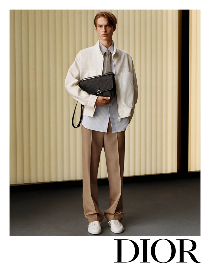 Model wearing a white jacket and beige trousers holding a black messenger bag in a minimalist interior setting