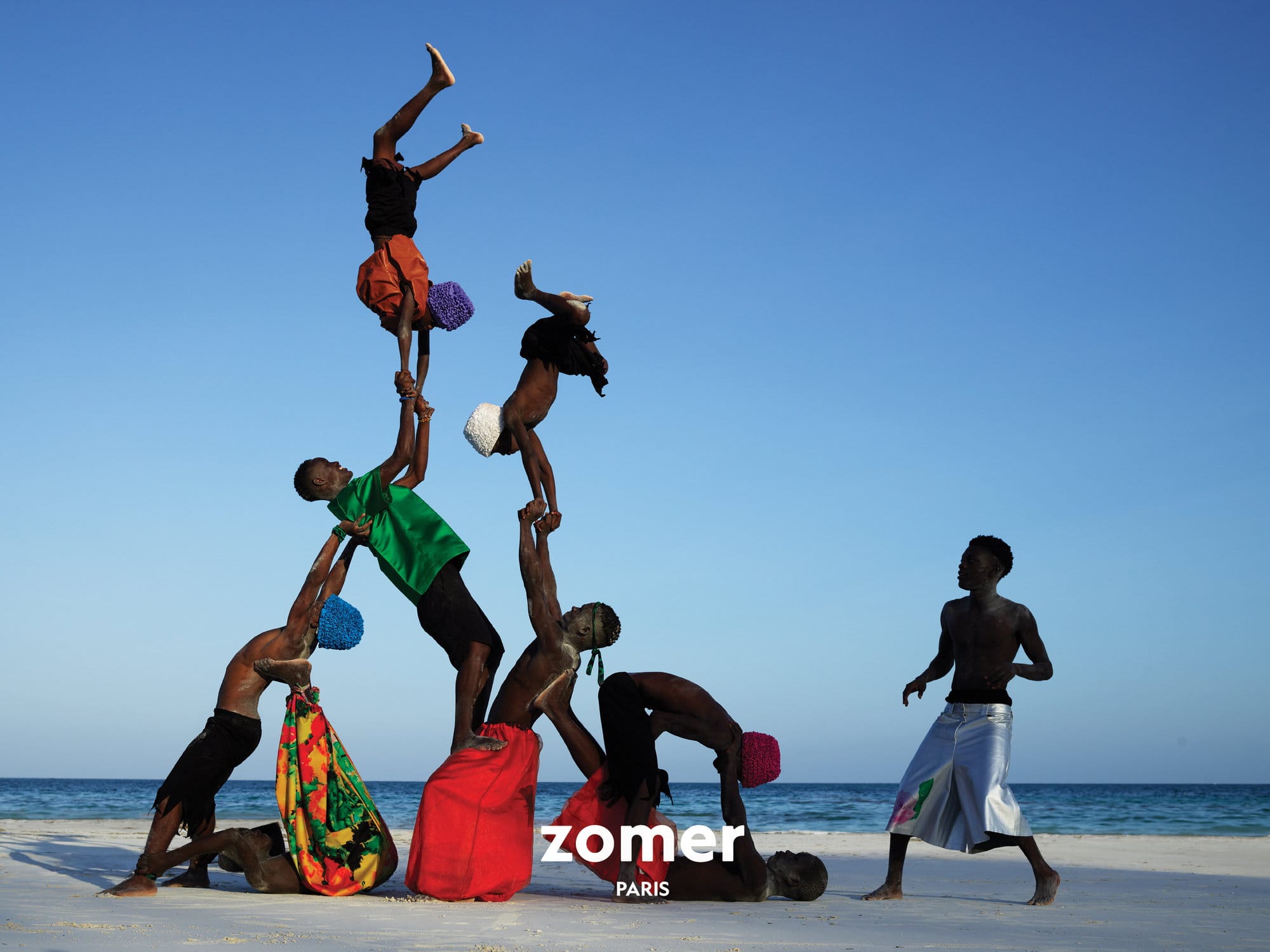 Group of acrobatic performers in a human pyramid formation on a white sandy beach against a clear blue sky, for Zomer.