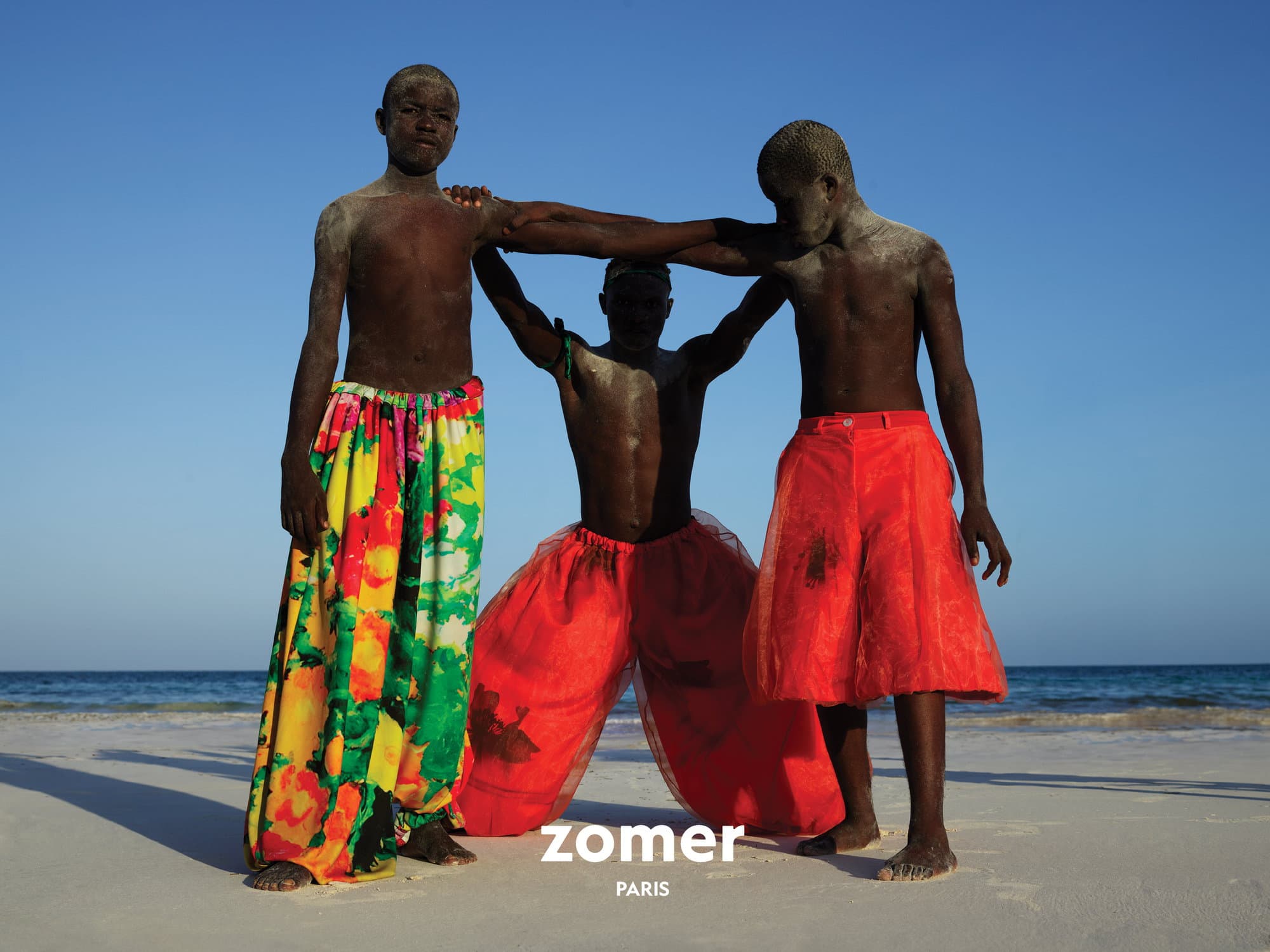 Three models standing on a sandy beach under a clear blue sky, wearing brightly colored trousers and shorts.