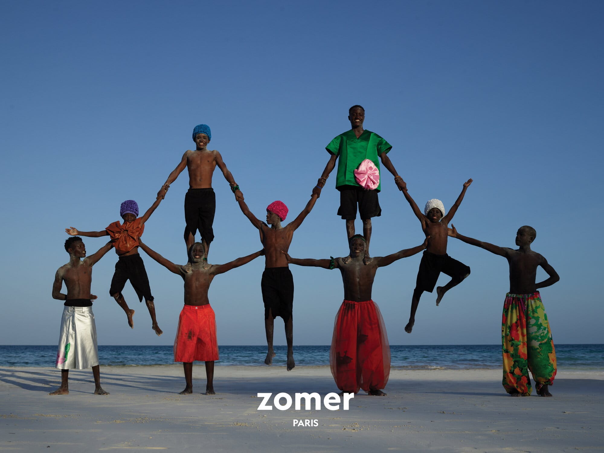 Group of models forming a human pyramid formation on a sandy beach by the ocean for Zomer Spring 2025 ad campaign.