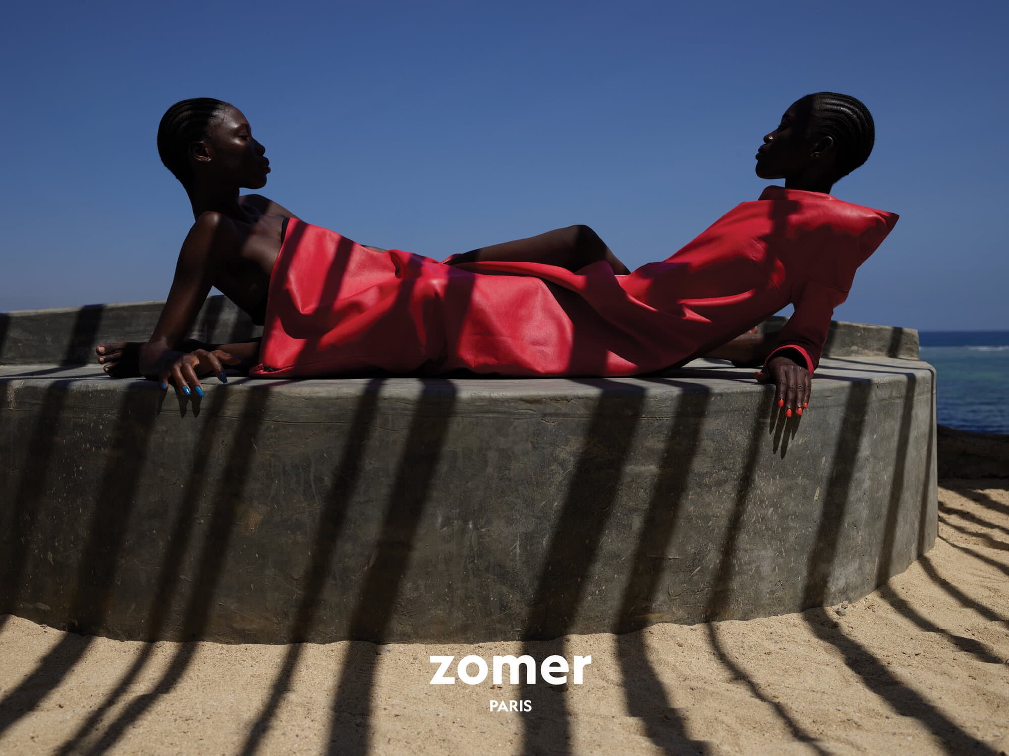 Two black female models in bright red dresses reclining on a curved concrete structure against a blue sky and ocean background.