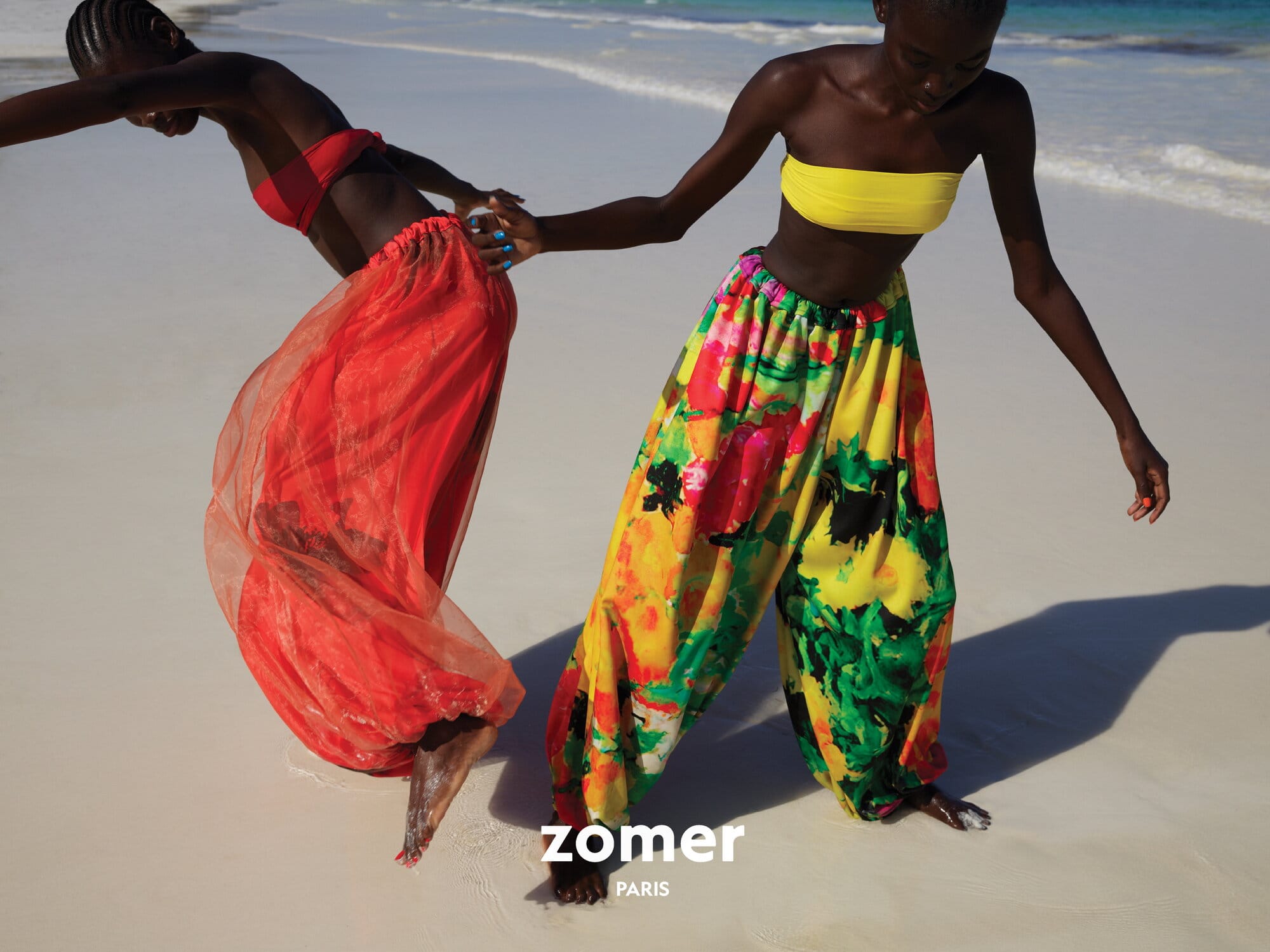 Two Black female models wearing vibrant swimwear walking on a sandy beach for Zomer.