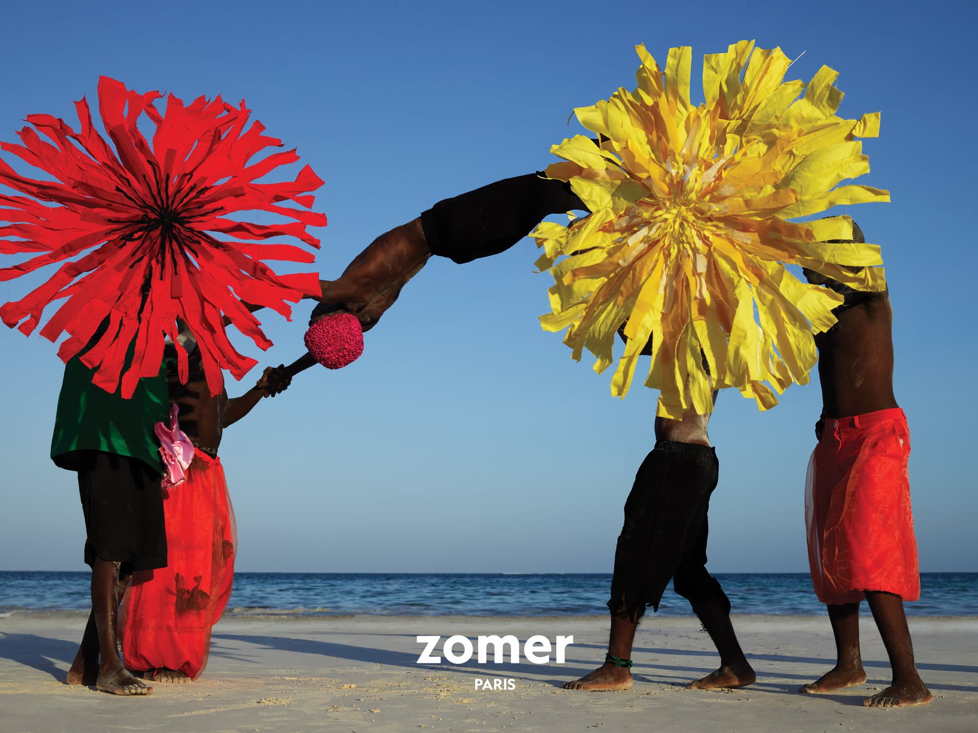 People on a sandy beach holding large paper flower props against a clear blue sky and ocean backdrop for a Zomer ad campaign.