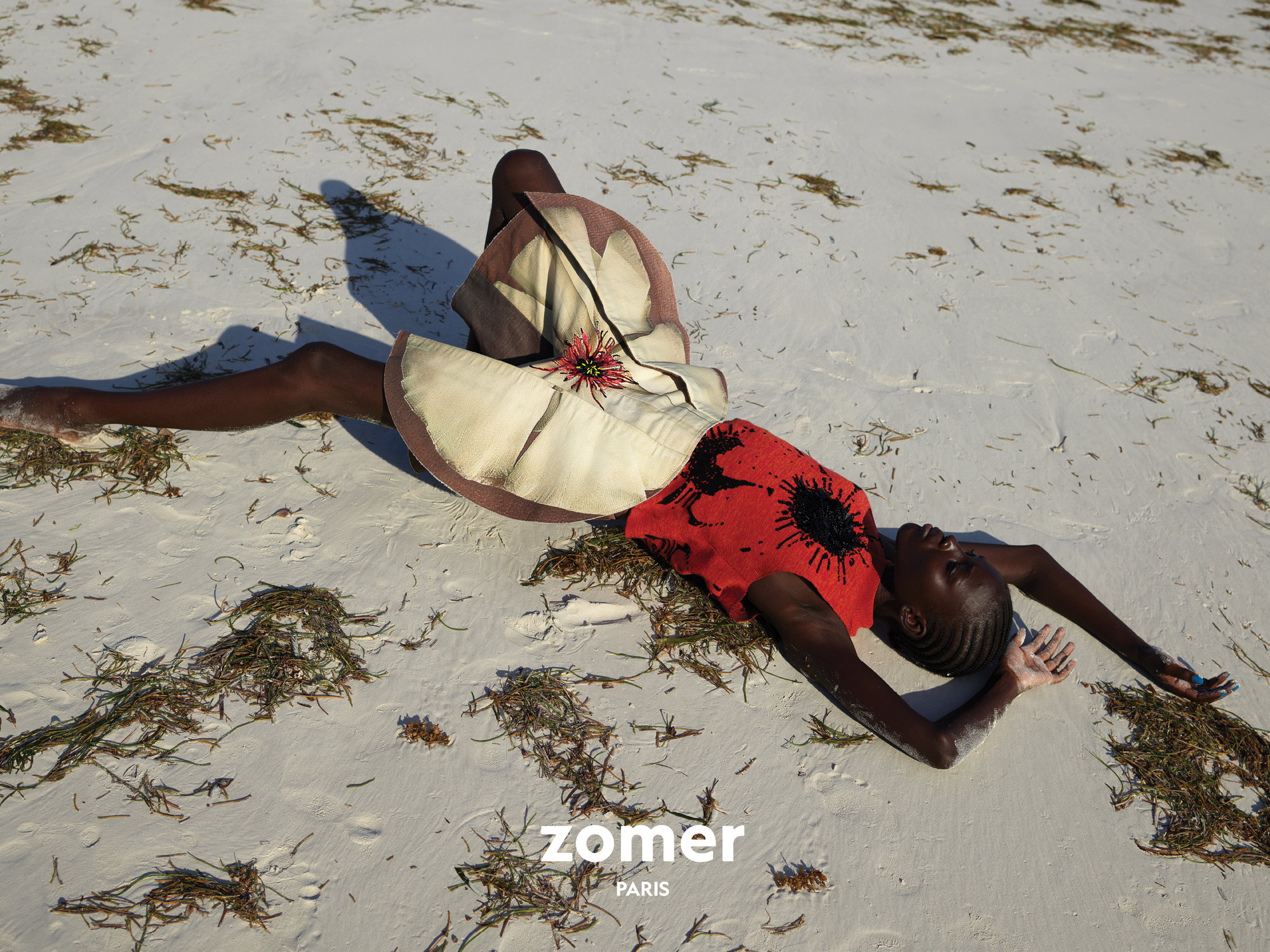 Model lying on white sandy beach wearing a structured floral skirt and red top with dark abstract prints