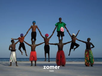 Group of models forming a human pyramid formation on a sandy beach by the ocean for Zomer Spring 2025 ad campaign.