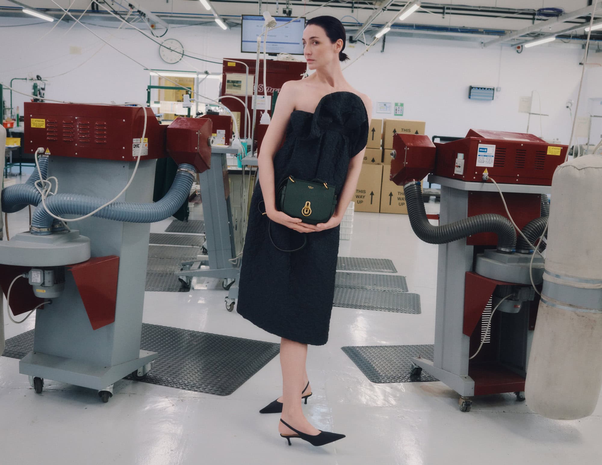 Model wearing a black strapless dress and holding a green shoulder bag inside a factory setting with industrial machinery