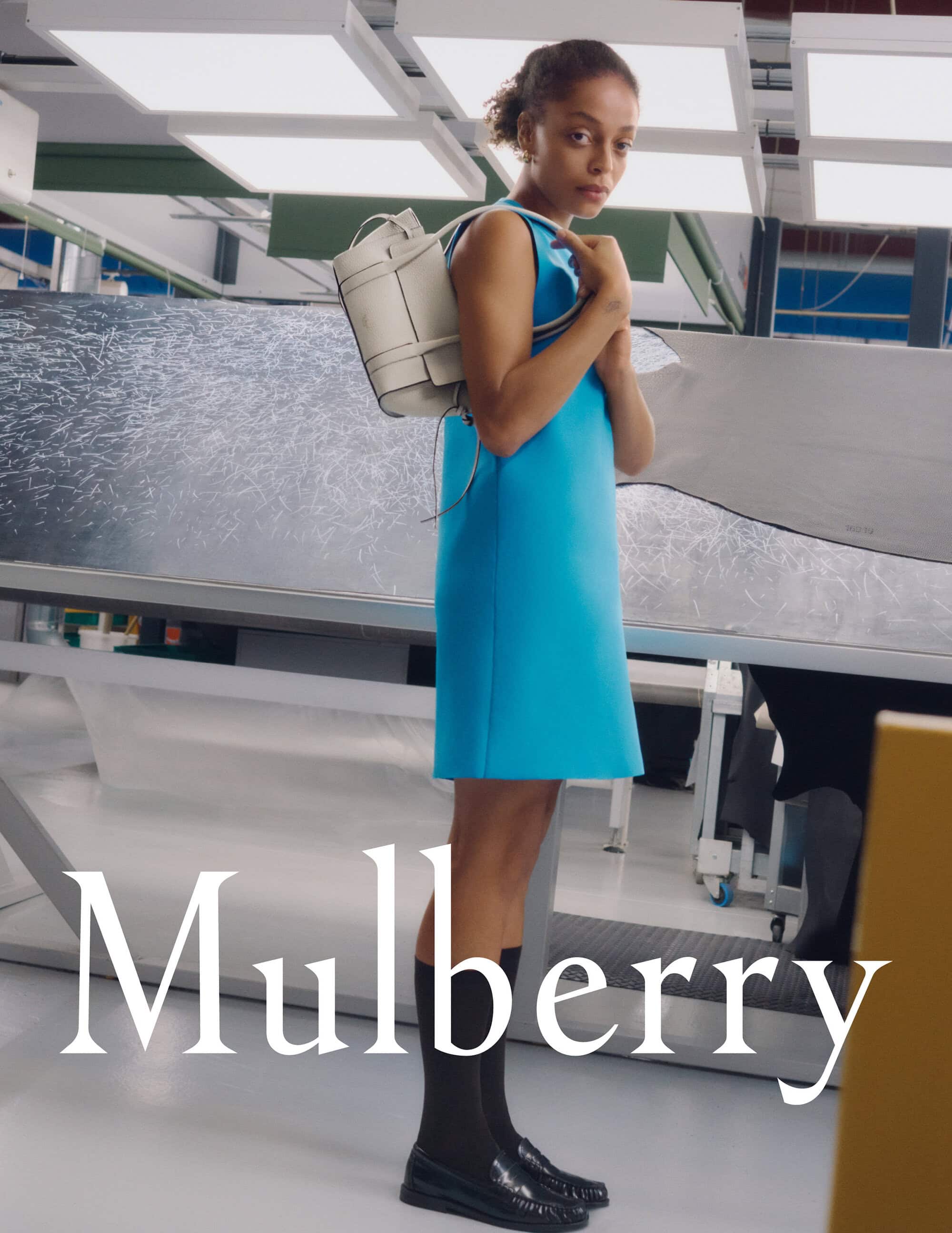 Model in a bright blue dress wearing a white leather backpack in an industrial factory interior for Mulberry