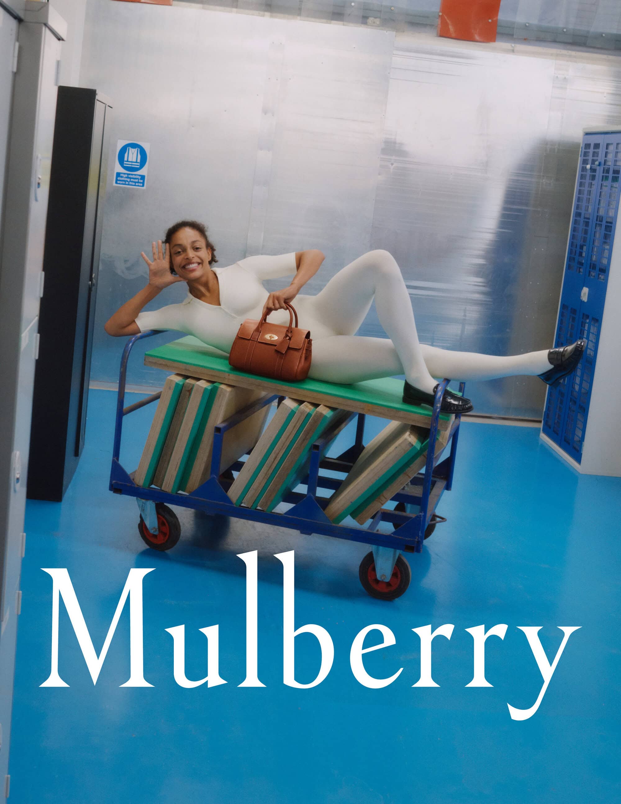 Model in white jumpsuit lying on a rolling utility cart holding a brown leather handbag in an industrial setting.
