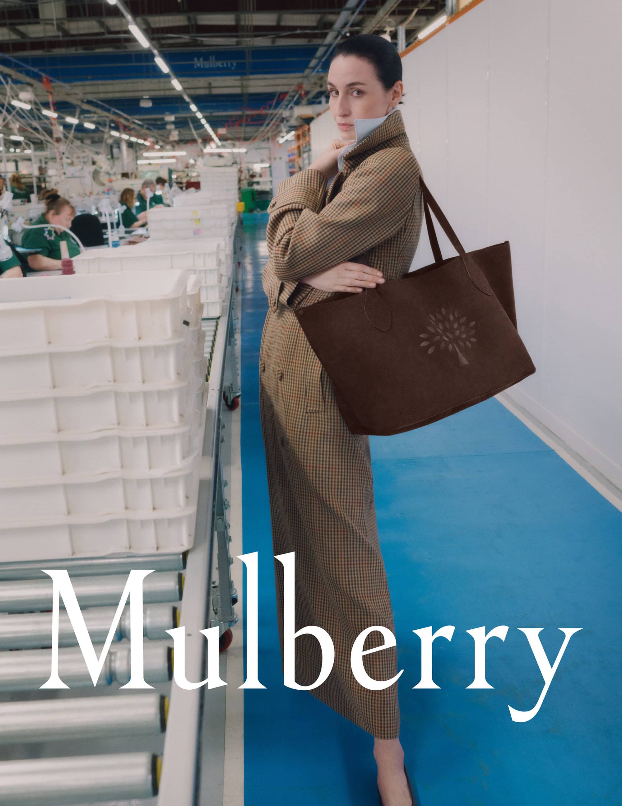 Model wearing a brown plaid coat holding a large brown tote bag inside a manufacturing facility with white assembly bins