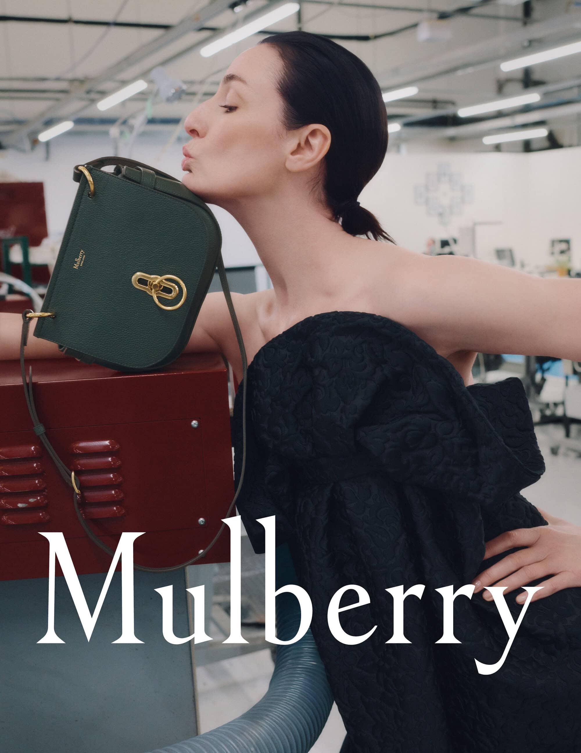 Model wearing dark textured dress leaning against red surface and kissing a green leather handbag