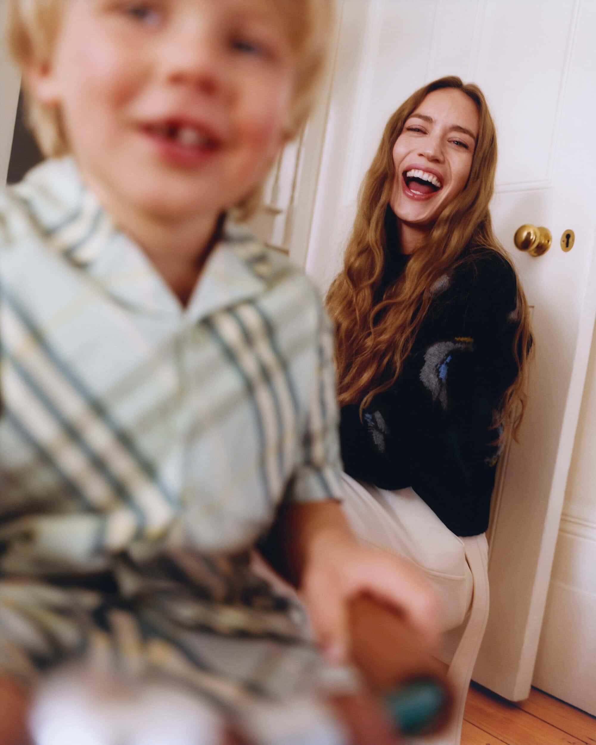 Laughing woman in dark sweater seated near a door with a blurry child in the foreground for Burberry Mother's Day 2025 ad campaign