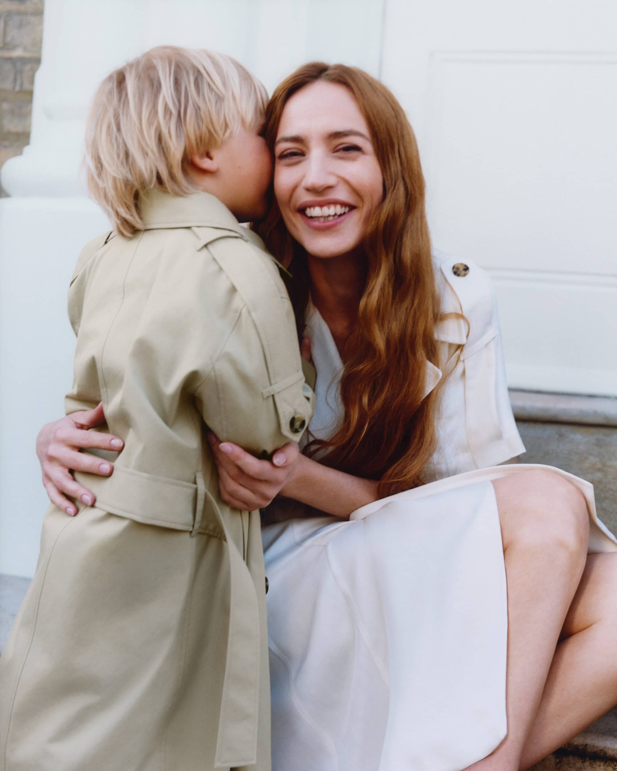 Woman smiling and embracing a child wearing a tan trench coat against a white background