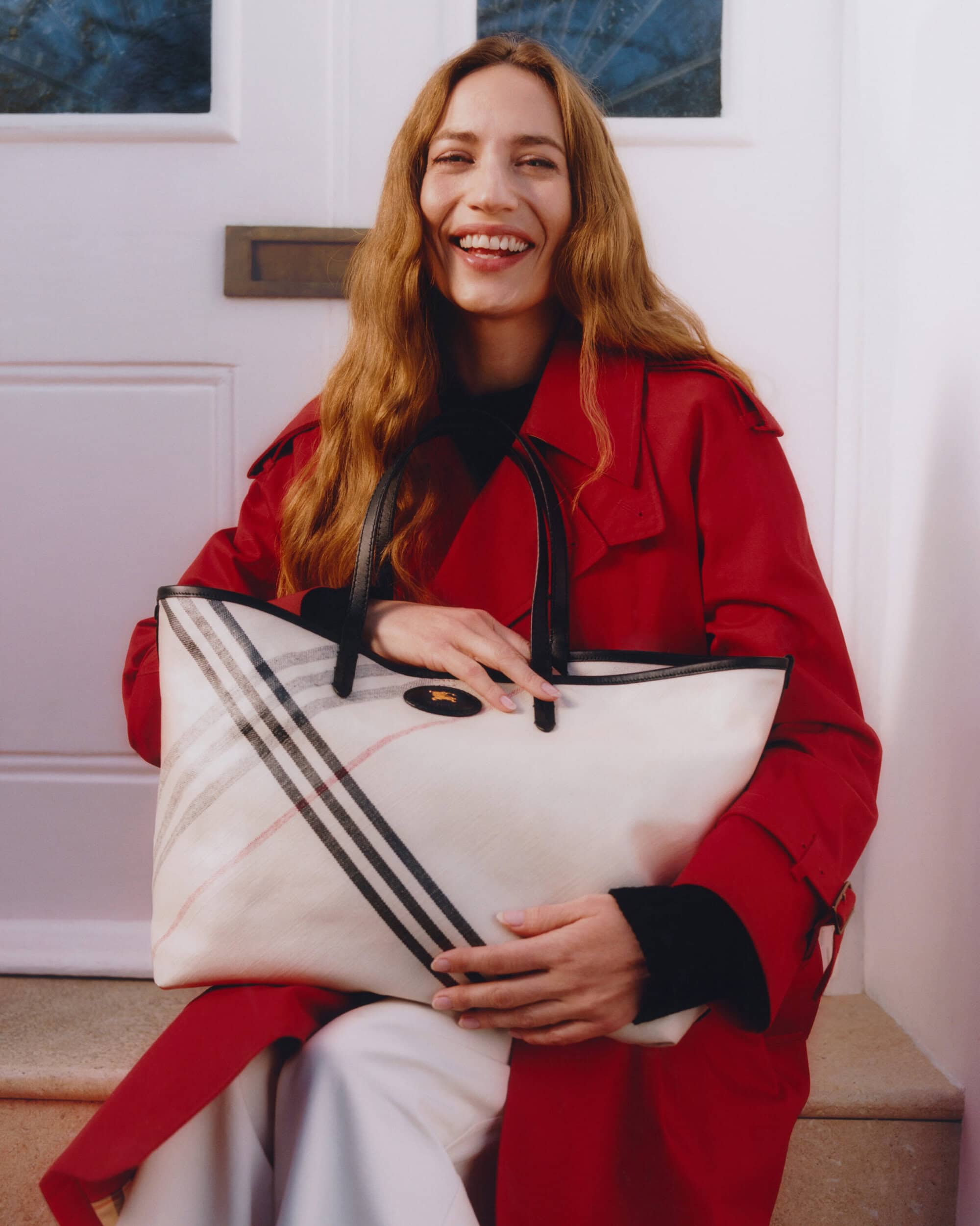 Smiling woman in red coat holding a large plaid tote bag seated on stone steps outside a white door