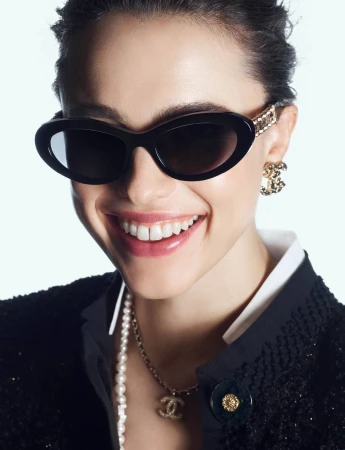 Close-up portrait of a smiling woman wearing black sunglasses, pearl necklace, and black textured jacket against a white background.
