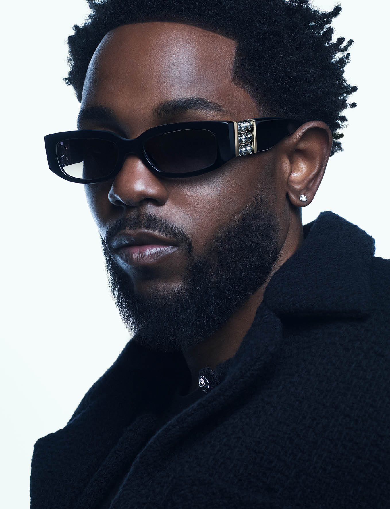 Close-up portrait of Kendrick Lamar wearing black sunglasses and a dark textured jacket against a white background.