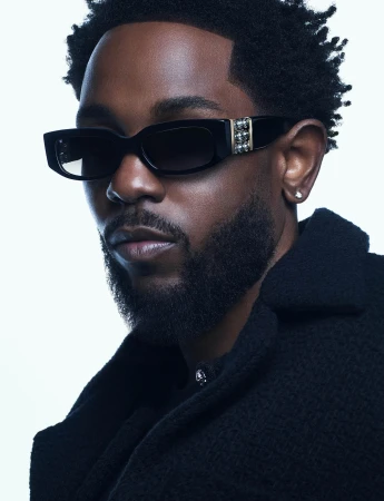 Close-up portrait of Kendrick Lamar wearing black sunglasses and a dark textured jacket against a white background.
