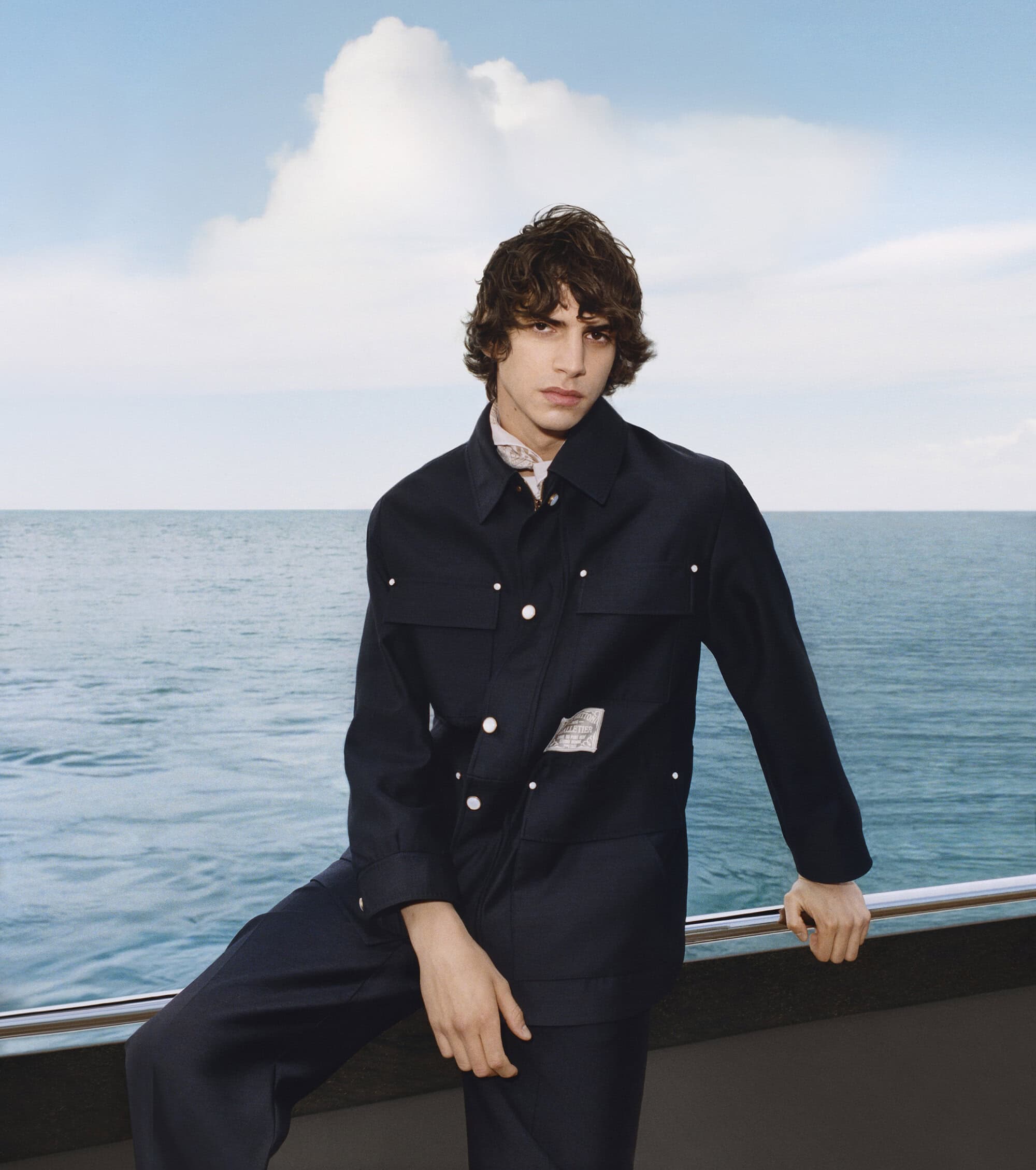 Young man in a dark navy jacket leaning against a metal railing outdoors with the ocean and sky in the background.