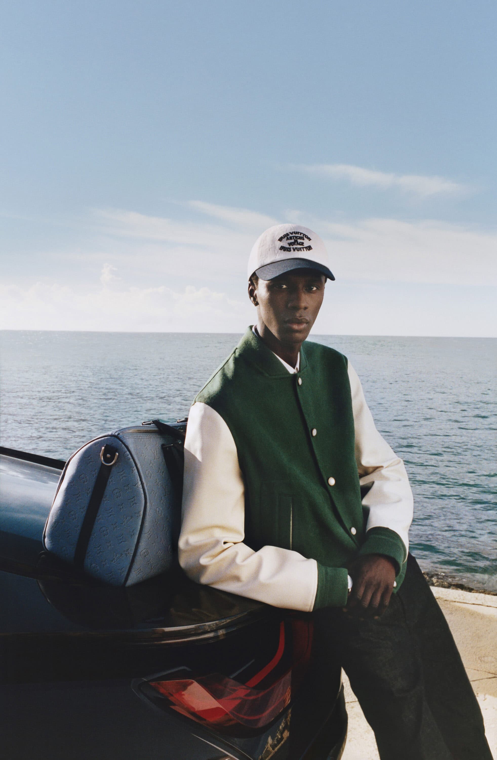 Black man wearing a green and white varsity jacket and baseball cap leaning against a car by the ocean.