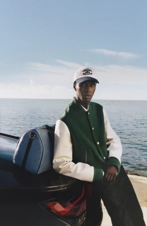 Black man wearing a green and white varsity jacket and baseball cap leaning against a car by the ocean.