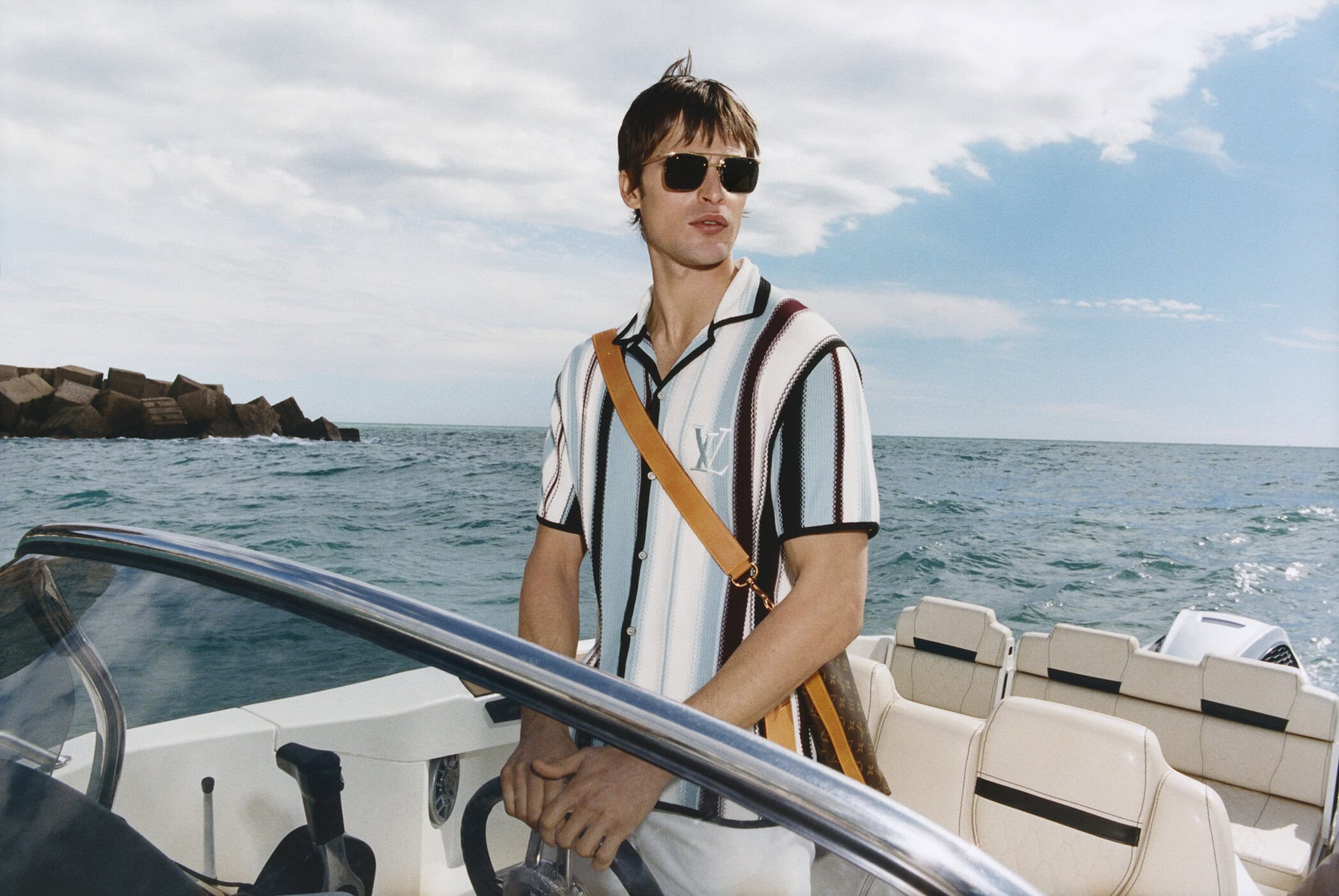 A man wearing sunglasses and a striped knit polo steering a motorboat on the ocean