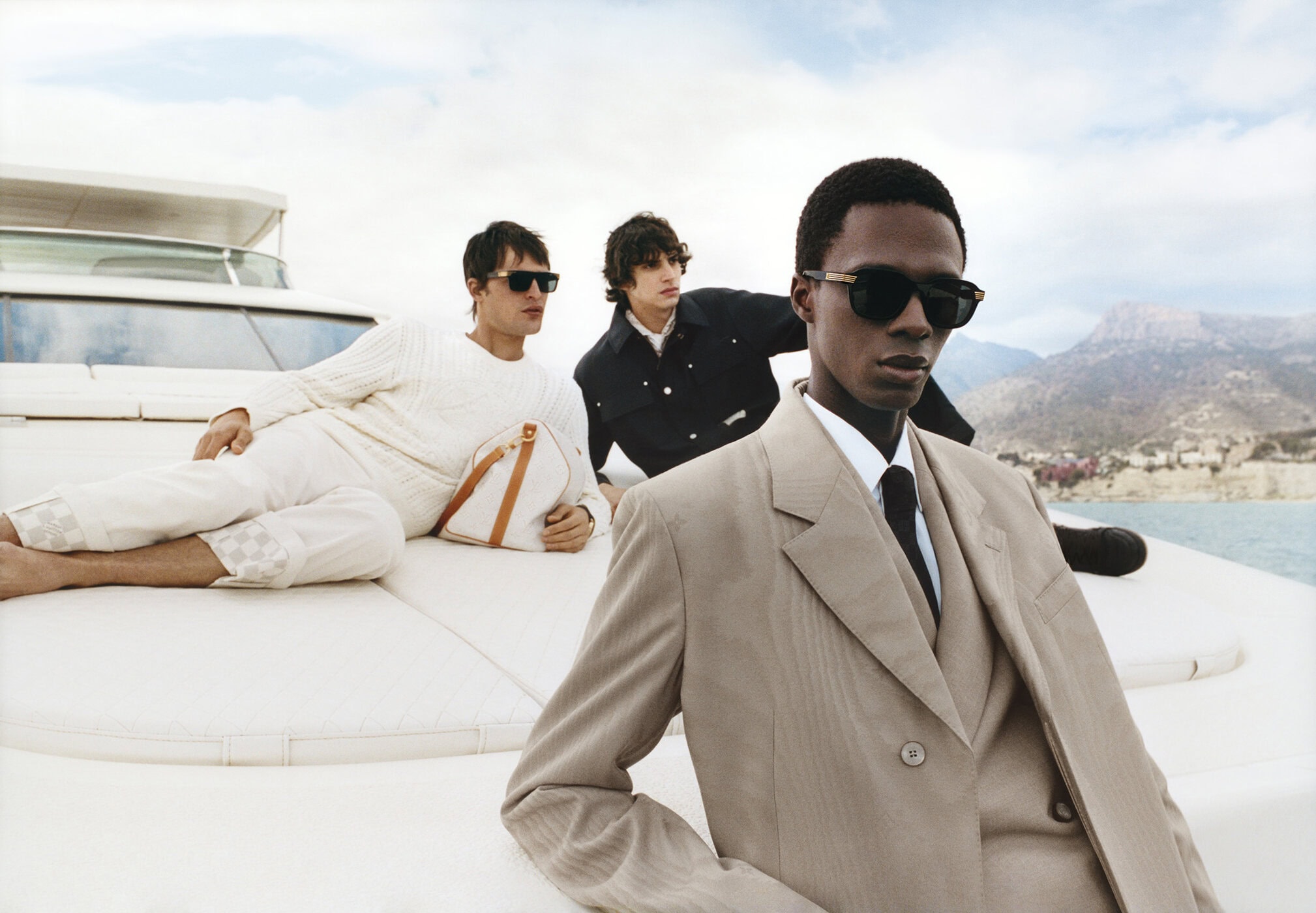 Three male models in menswear on a yacht deck, with a coastal mountain background for Louis Vuitton Pre-Fall 2025.