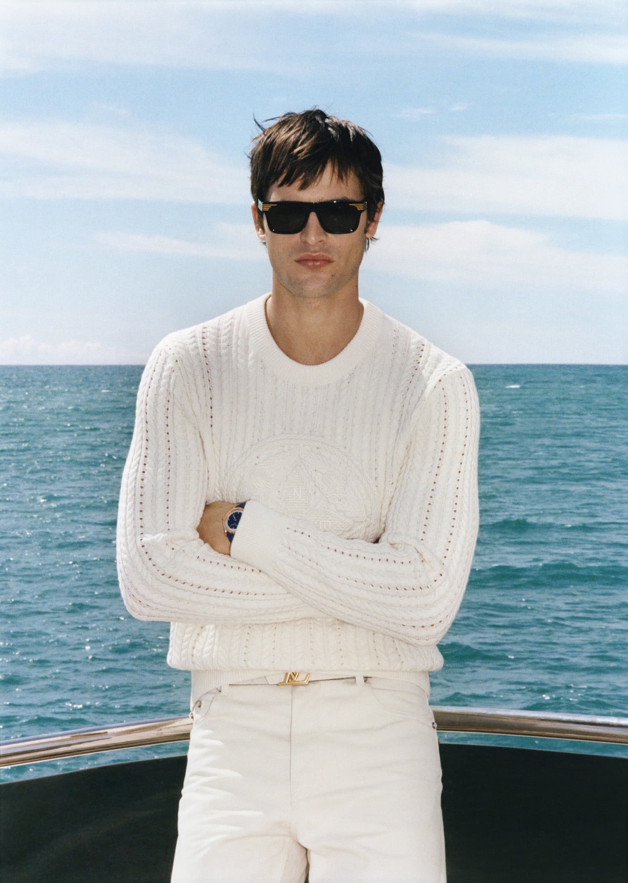 Man wearing a white knit sweater and sunglasses standing on a boat with ocean background