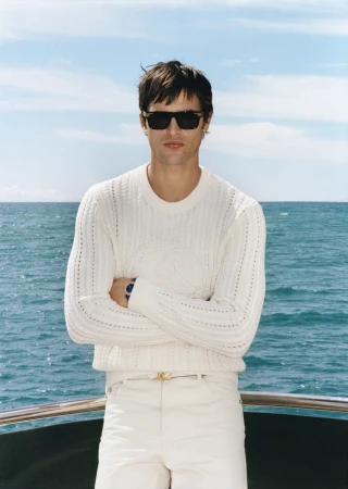 Man wearing a white knit sweater and sunglasses standing on a boat with ocean background