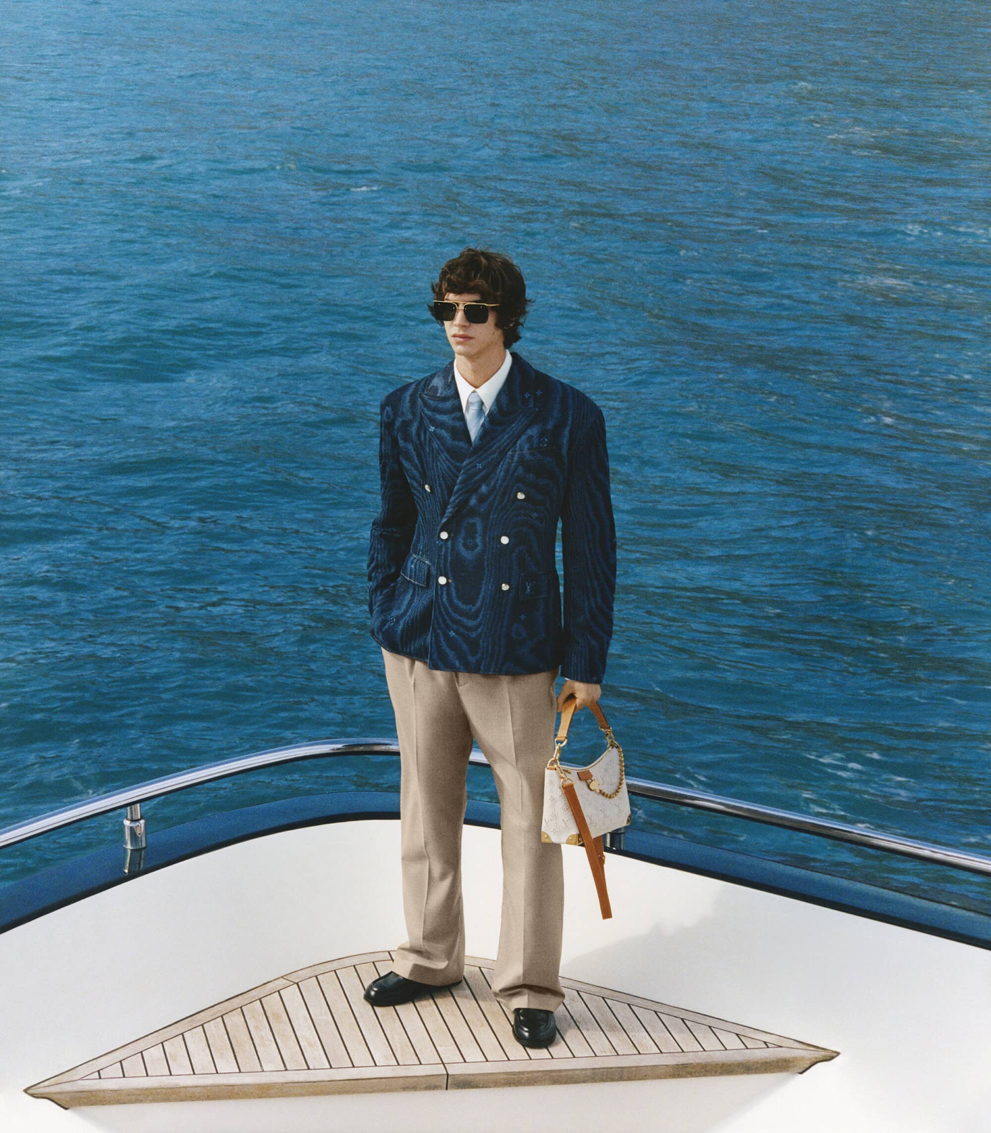 Man wearing a textured blue double-breasted blazer and holding a white handbag standing on a boat deck
