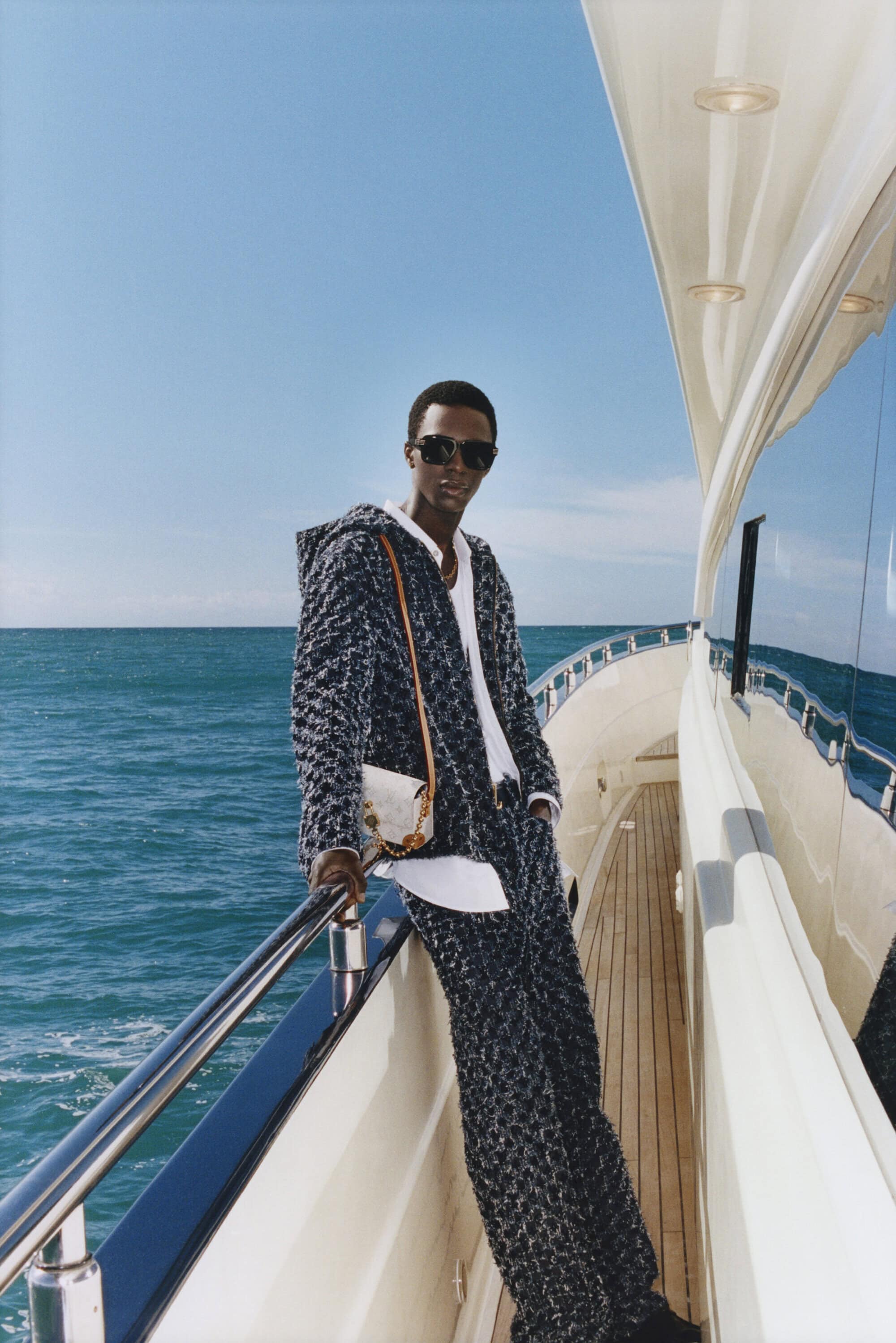 Man wearing sunglasses and patterned hoodie leaning on a yacht railing against the ocean
