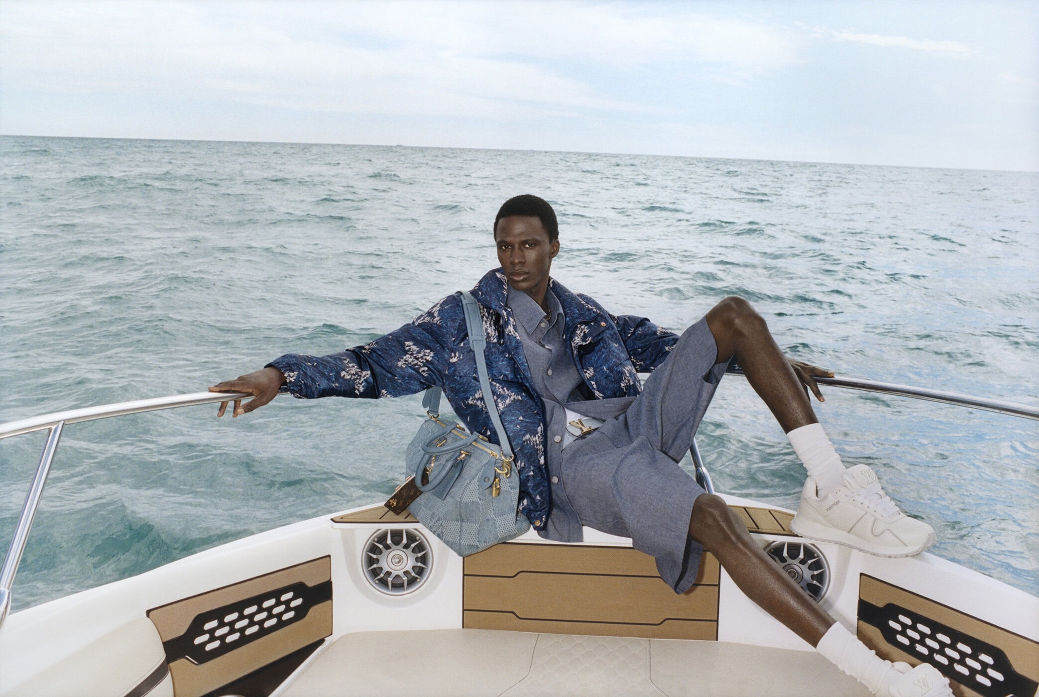 Black man seated on a boat railing wearing a blue printed jacket, shorts, and holding a light blue handbag against the ocean.