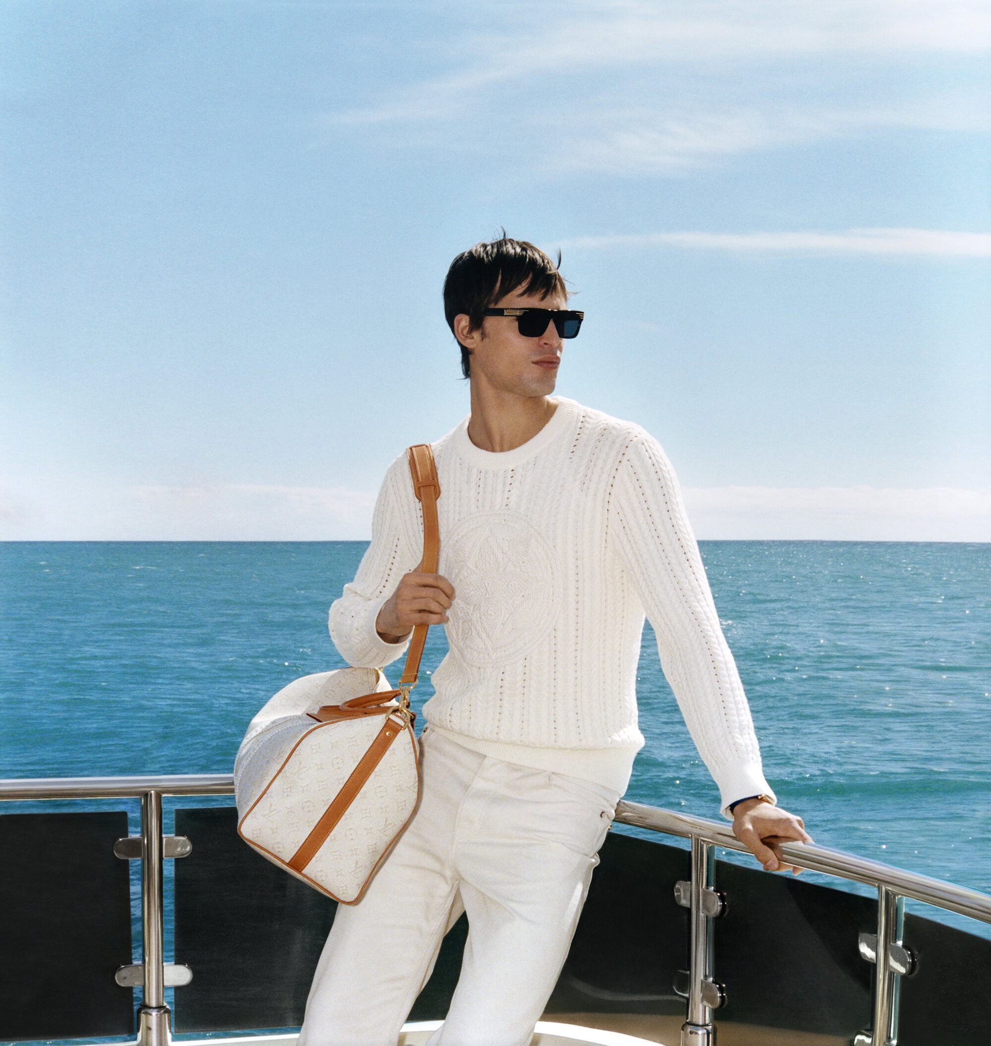 Man wearing a white knit sweater and sunglasses leaning on a metal railing with the ocean in the background