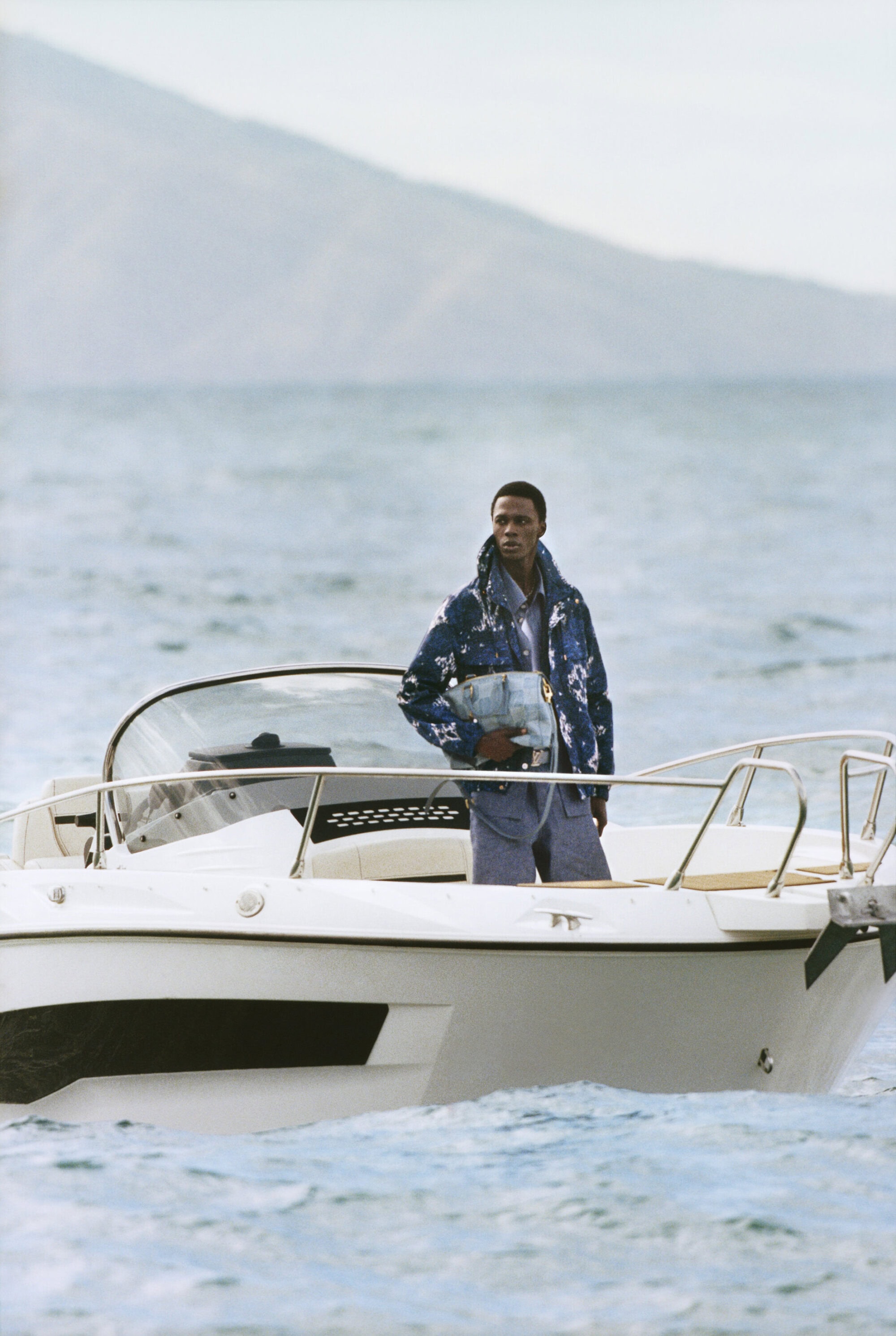 Man wearing a blue patterned jacket holding a light blue handbag aboard a white motorboat on the ocean