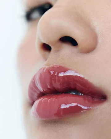 Close-up on a woman's face featuring glossy red lips, a nose, and a blurred eye against a white background for beauty and fashion.