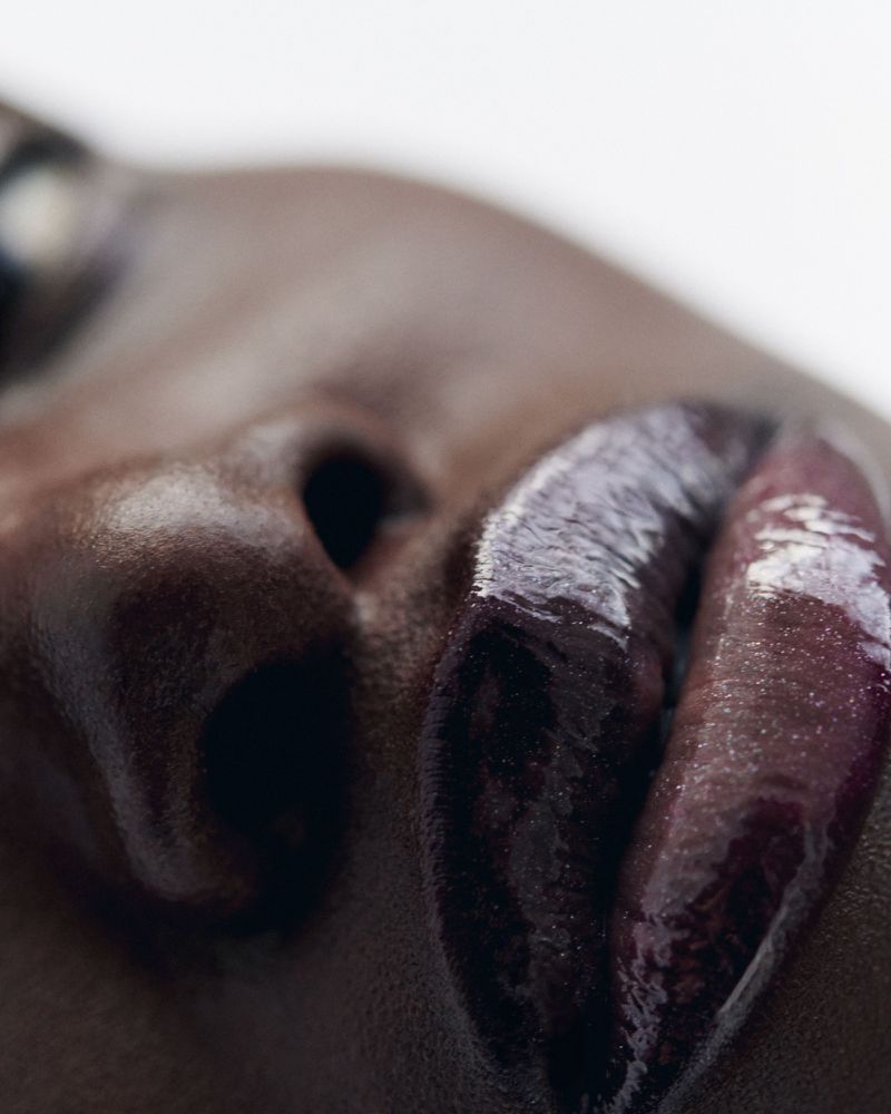Close-up shot of an adult woman's face with dark skin and glossy purple lipstick, focusing on the lips and nose against a white background.